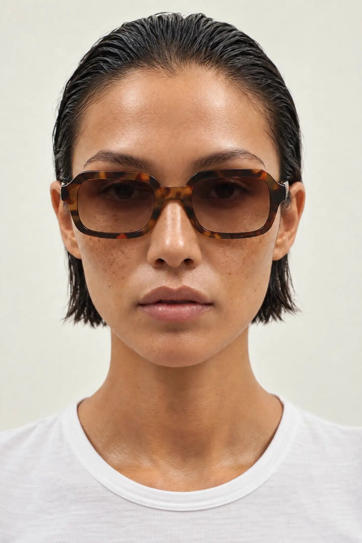 A close-up portrait of a model wearing oversized square sunglasses with a tortoiseshell pattern and dark tinted lenses, paired with a white crew-neck t-shirt against a light beige background.