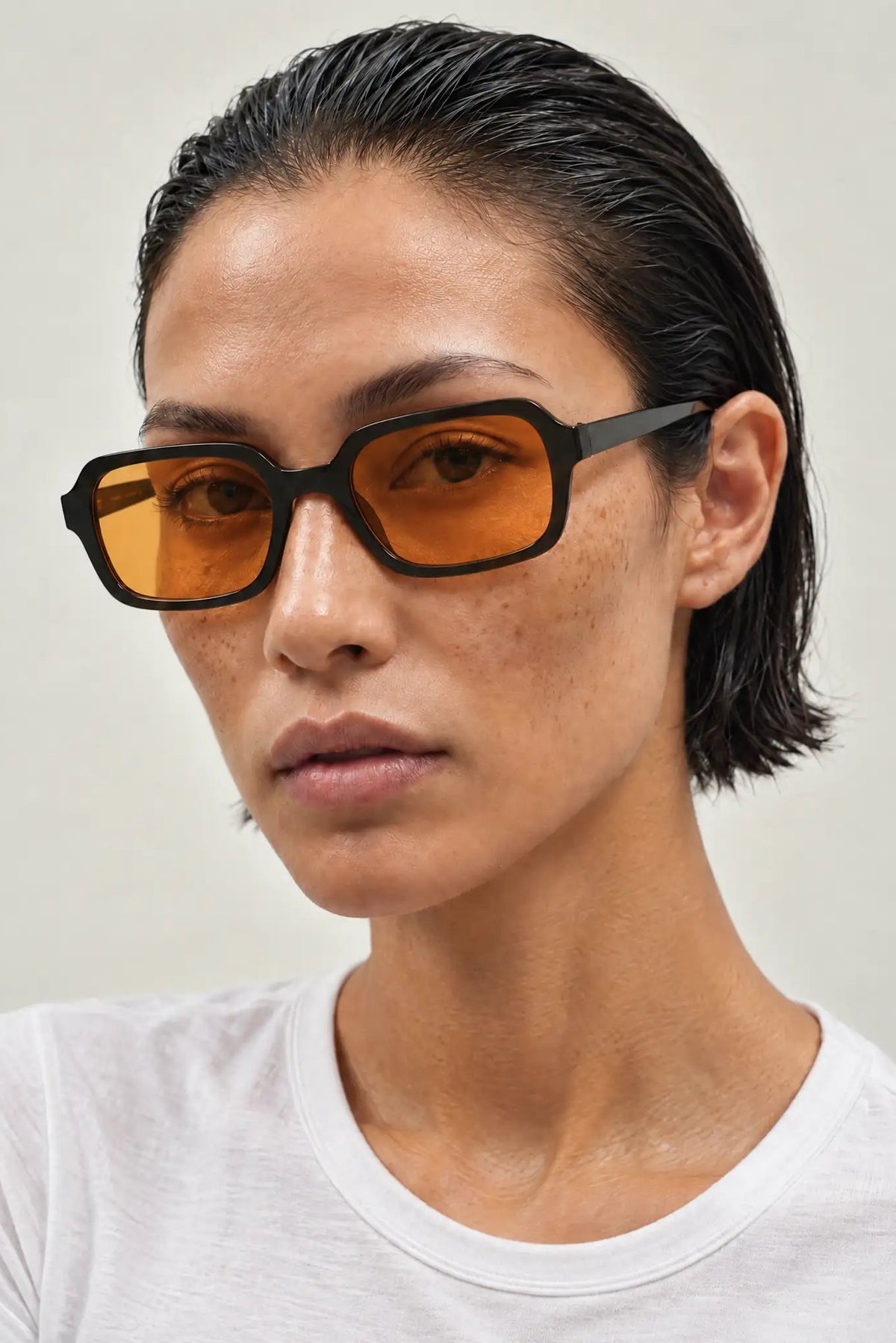 A close-up of a model wearing thick black square sunglasses with amber-orange tinted lenses and a white t-shirt against a light gray background.