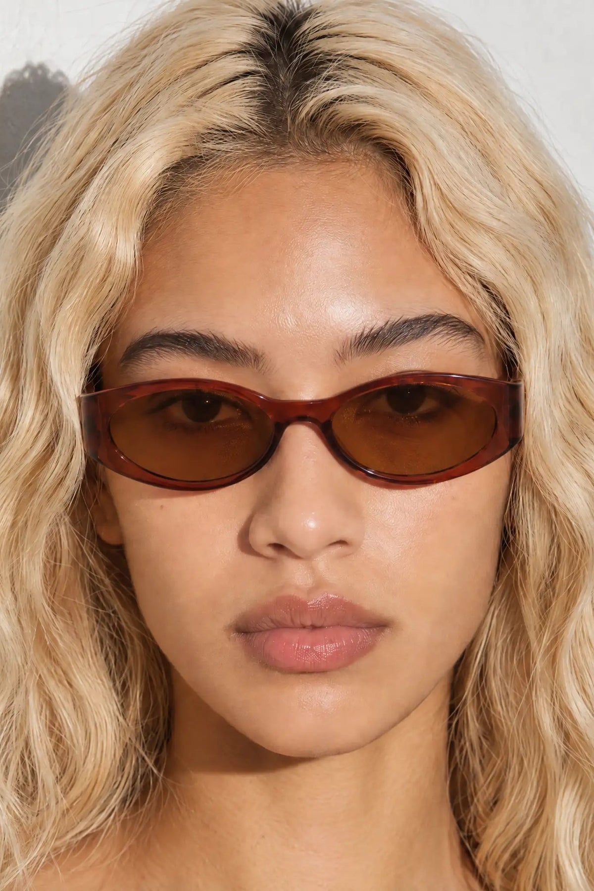 A close-up of a model with blonde wavy hair wearing slim, cat-eye style sunglasses with a translucent reddish-brown frame and dark brown tinted lenses.