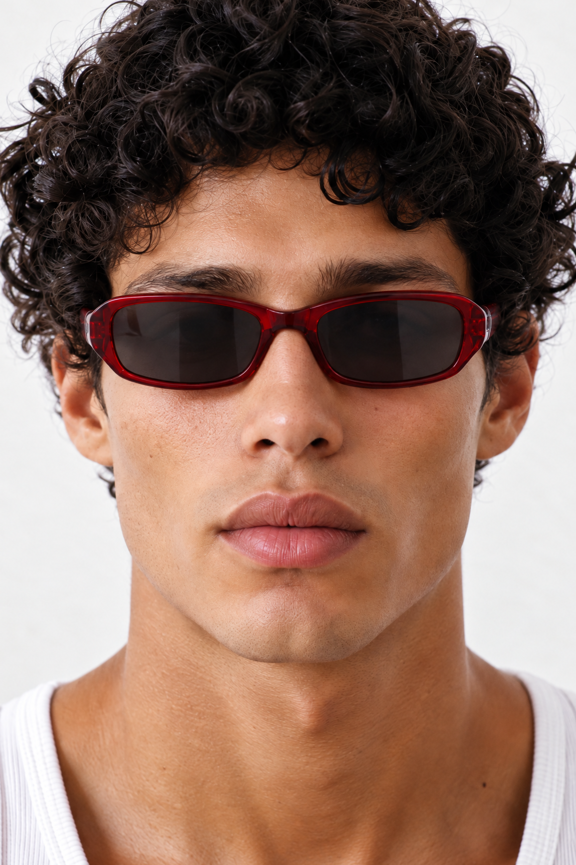 A close-up of a man with curly dark hair wearing RECTANGLE sunglasses with a translucent red frame and dark lenses, paired with a white ribbed tank top against a light gray background.