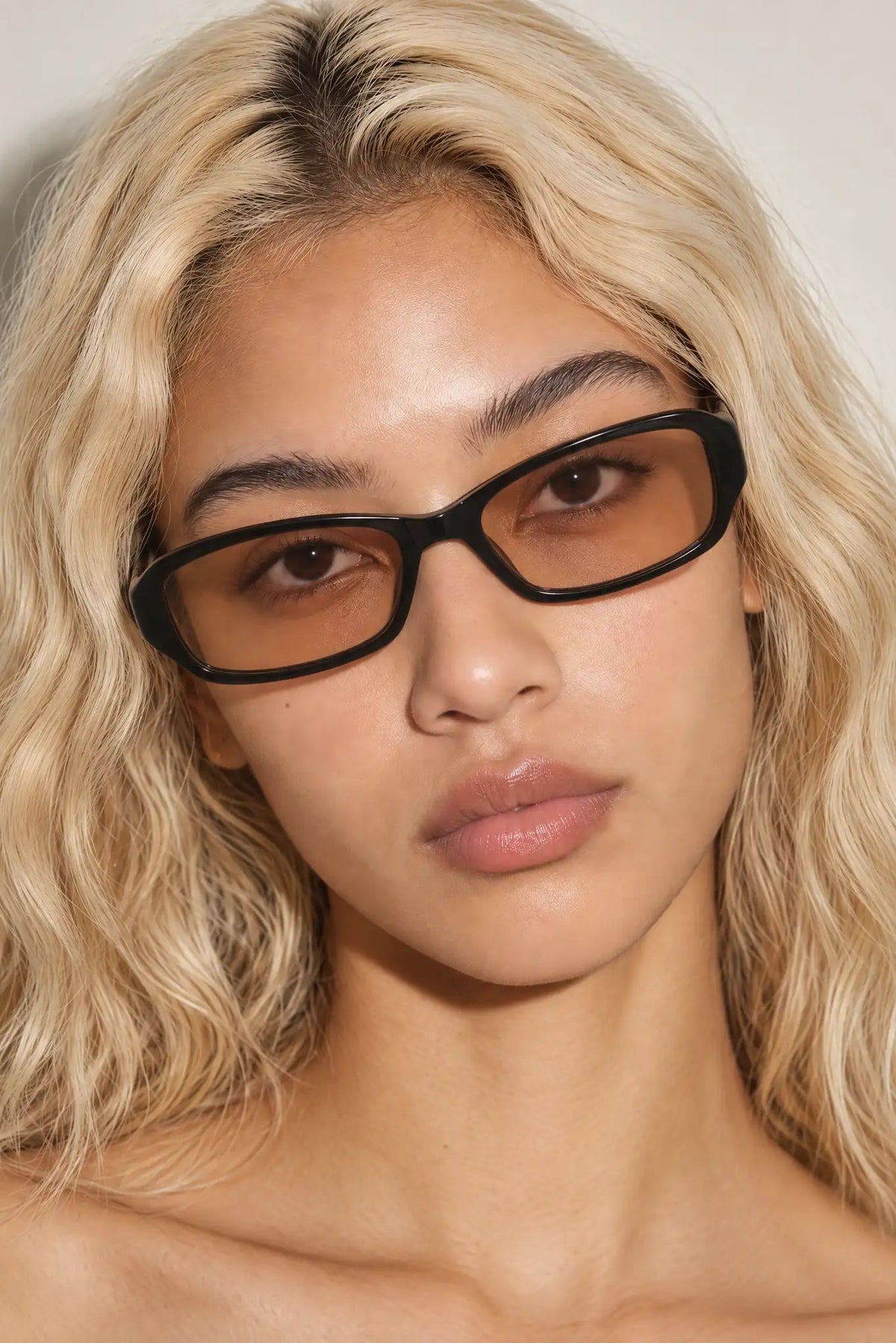 A close-up portrait of a woman with wavy blonde hair wearing thick black RECTANGLE eyeglasses with brown tinted lenses against a plain light background.