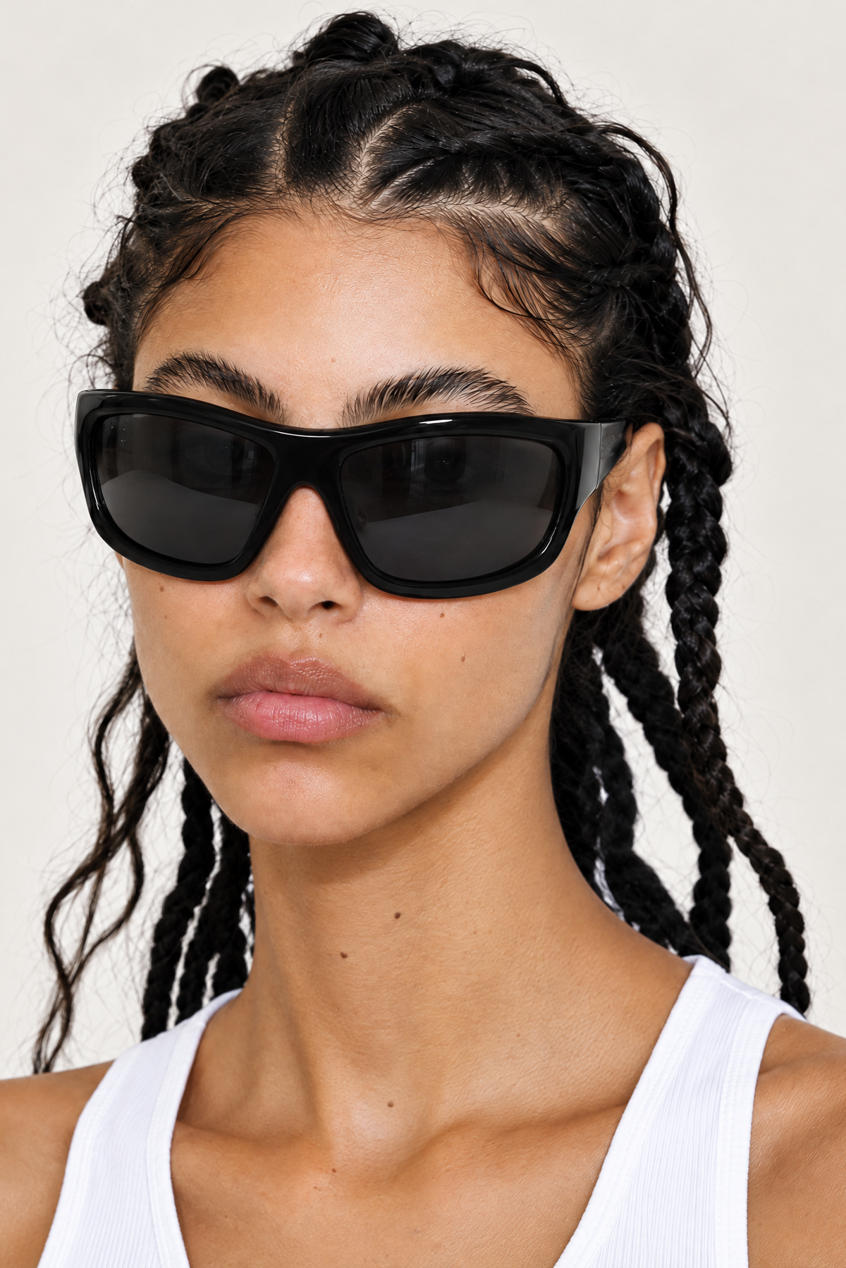 A close-up of a model with dark braided hair wearing oversized black sunglasses and a white ribbed tank top against a plain light background.