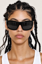 A close-up of a model with braided hair wearing thick black cat-eye sunglasses and a white ribbed tank top against a light beige background.