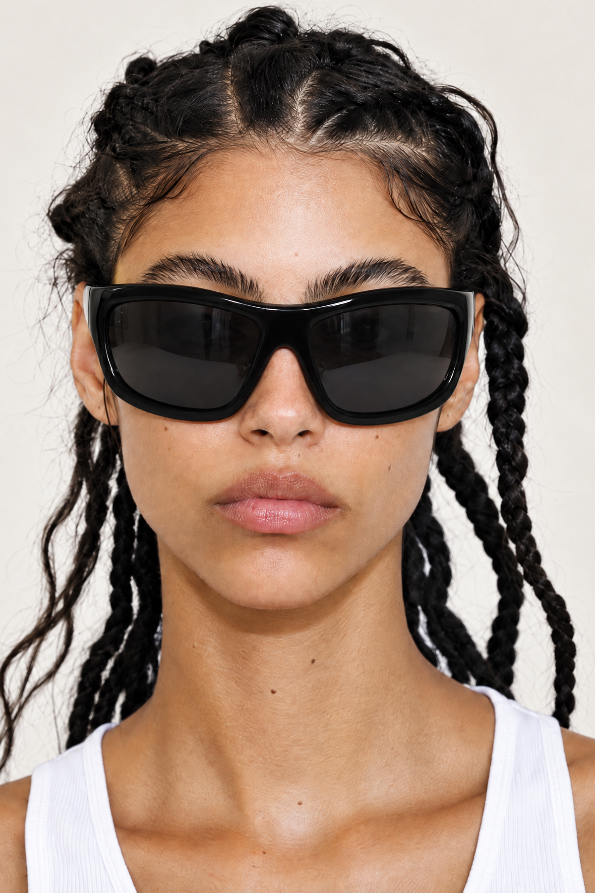 A close-up of a model with braided hair wearing thick black cat-eye sunglasses and a white ribbed tank top against a light beige background.