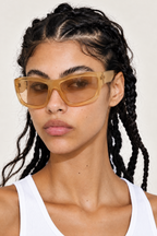 A close-up of a model with dark braided hair wearing oversized square sunglasses with a translucent amber-tinted frame and a white ribbed tank top, set against a plain light background.