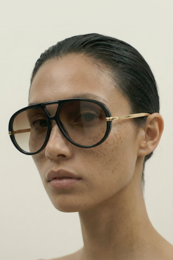 Person wearing black oversized aviator sunglasses with gold frames on a plain background