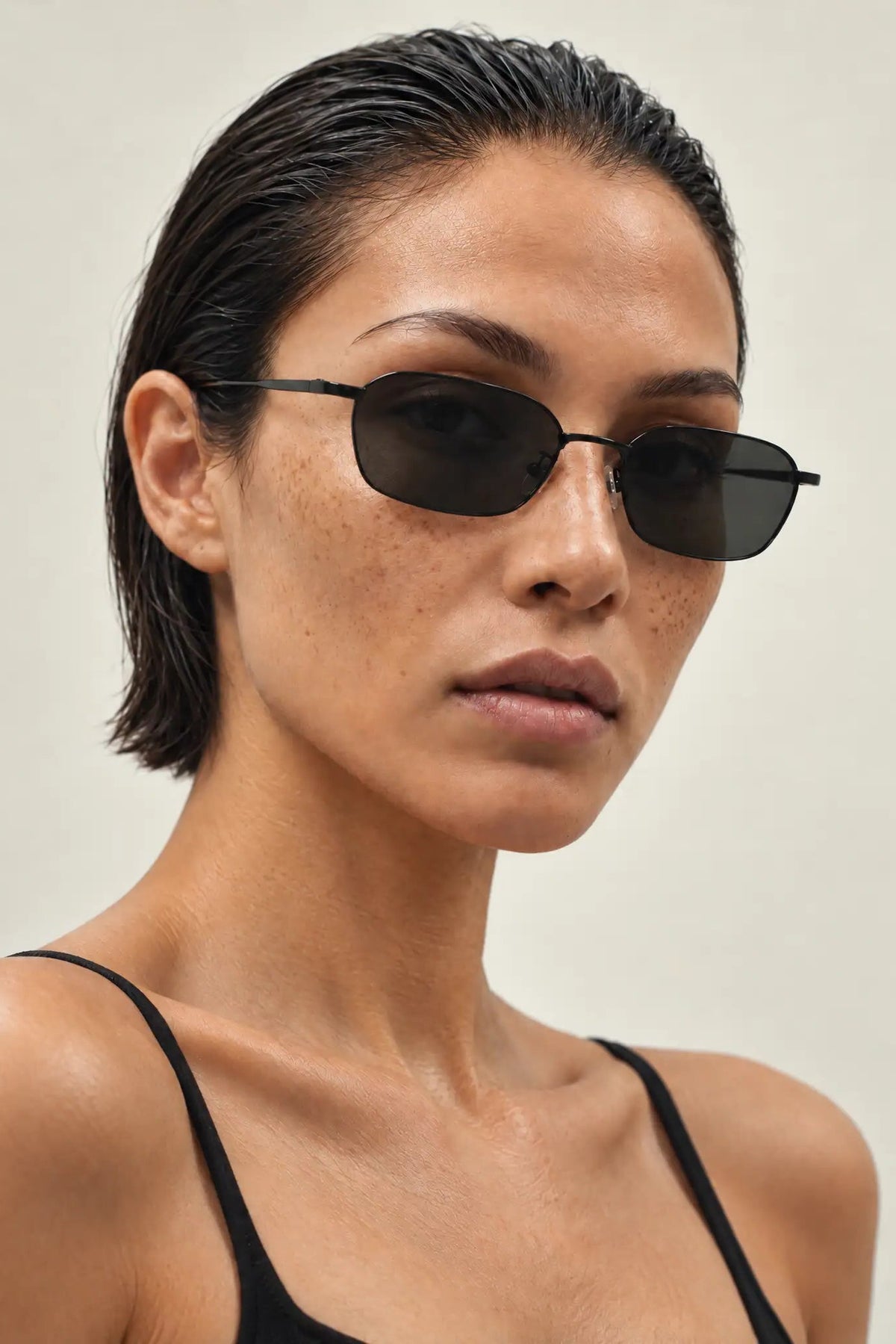 A close-up of a model with slicked-back dark hair wearing slim black metal oval sunglasses and a black spaghetti-strap top against a plain light beige background.