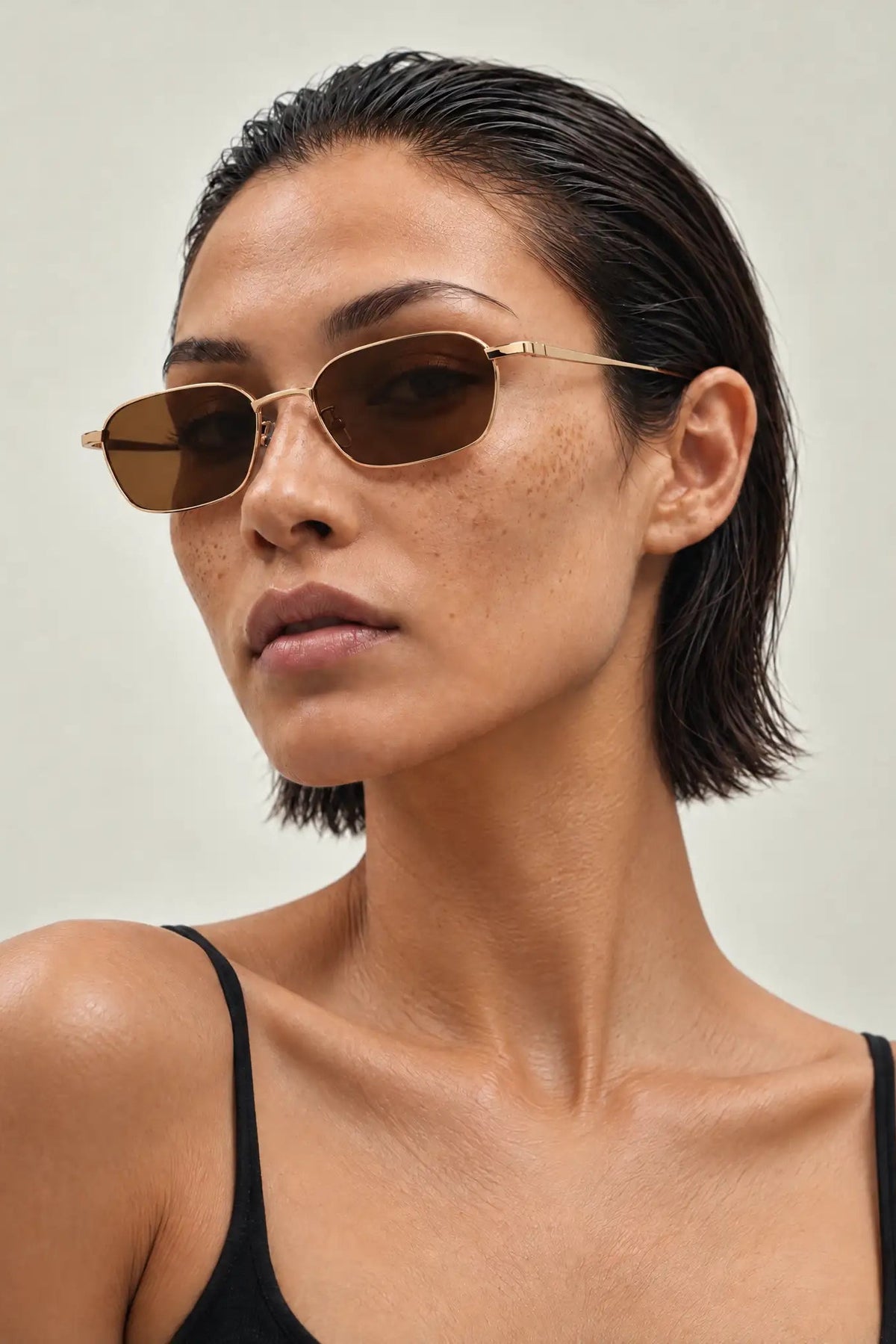 A close-up of a model wearing slim, angular gold-framed sunglasses with dark brown lenses, paired with a black spaghetti-strap top against a light beige background.