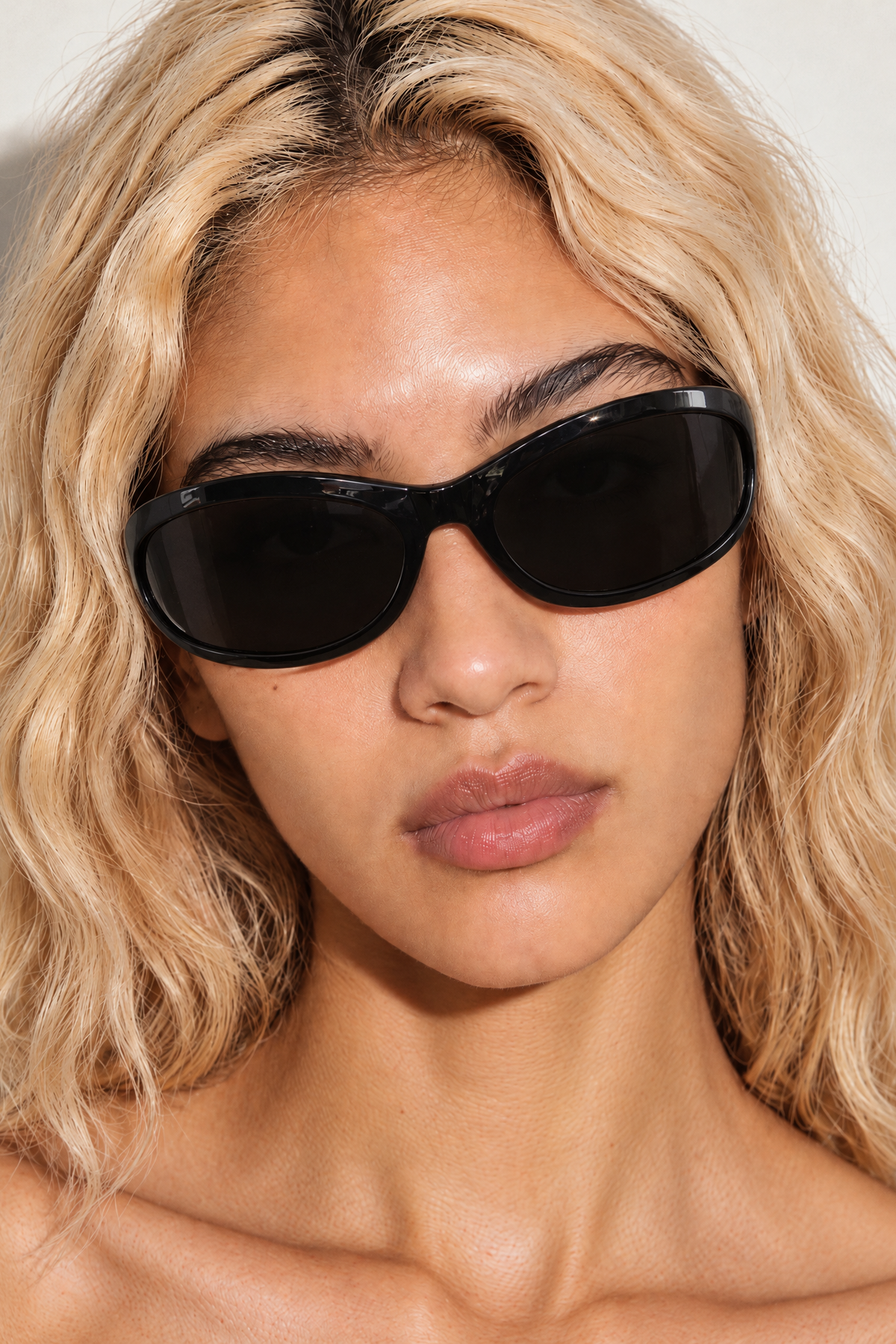A close-up of a model with wavy blonde hair wearing black wrap around sunglasses against a light neutral background.