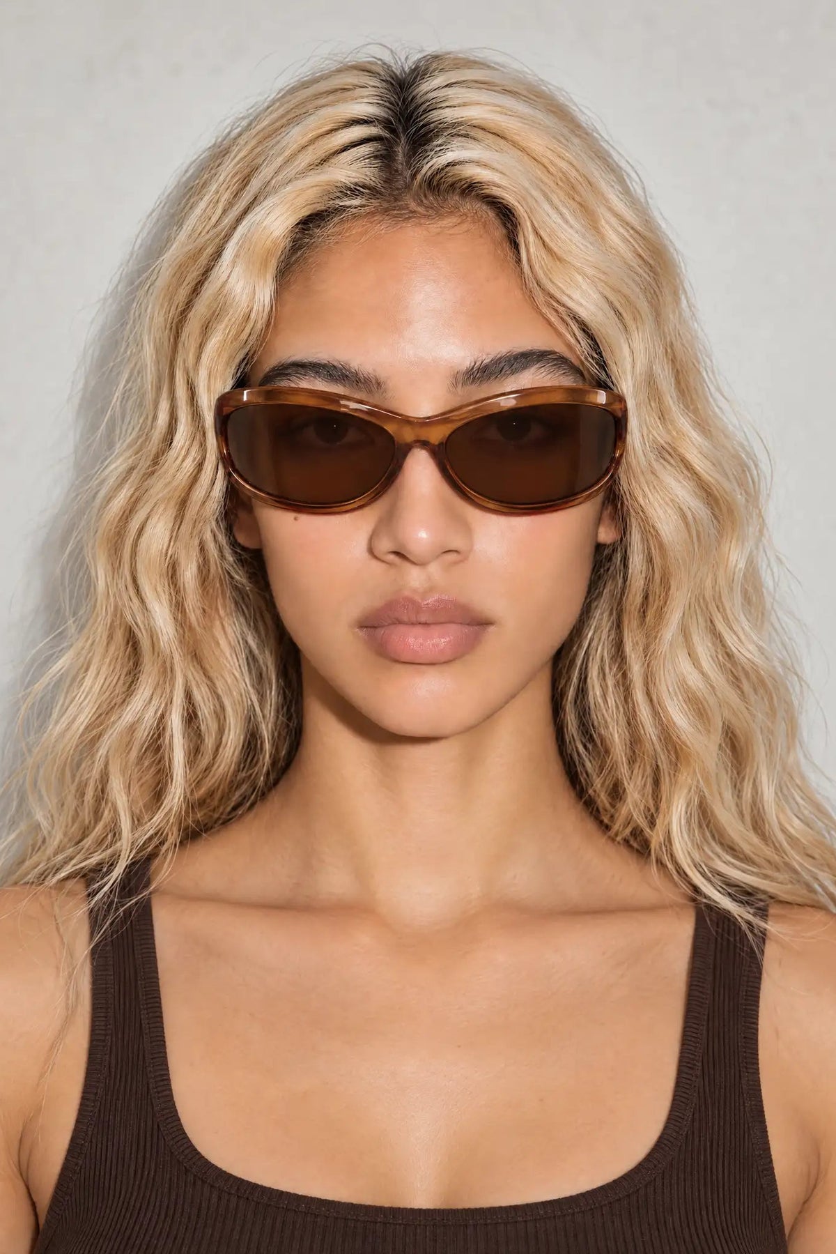 A woman with blonde wavy hair wearing wrap round sunglasses with brown frames and dark brown lenses, paired with a dark brown ribbed tank top against a light gray background.