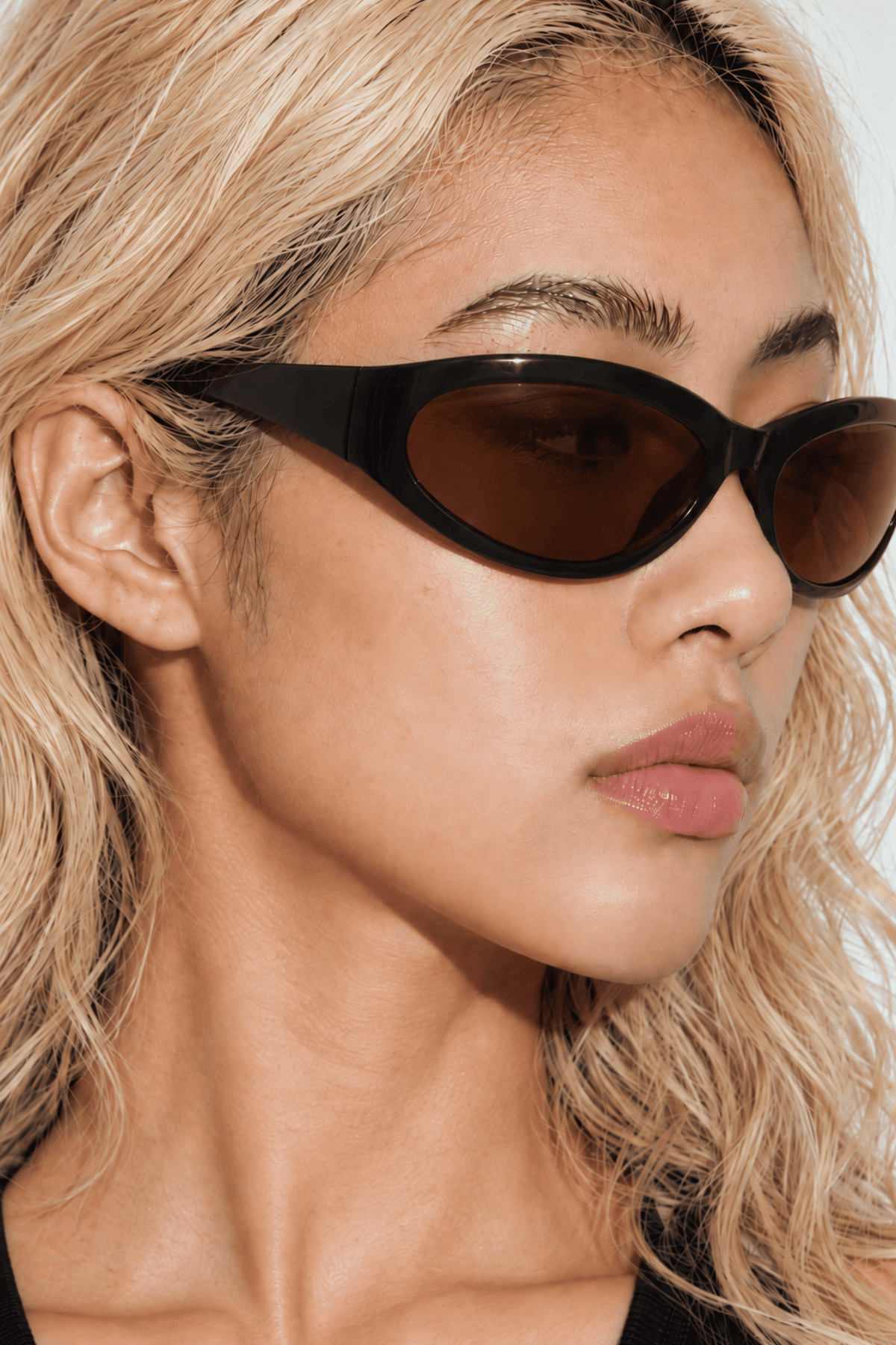 A close-up of a model with blonde wavy hair wearing black wrap around sunglasses with dark brown lenses and side profile