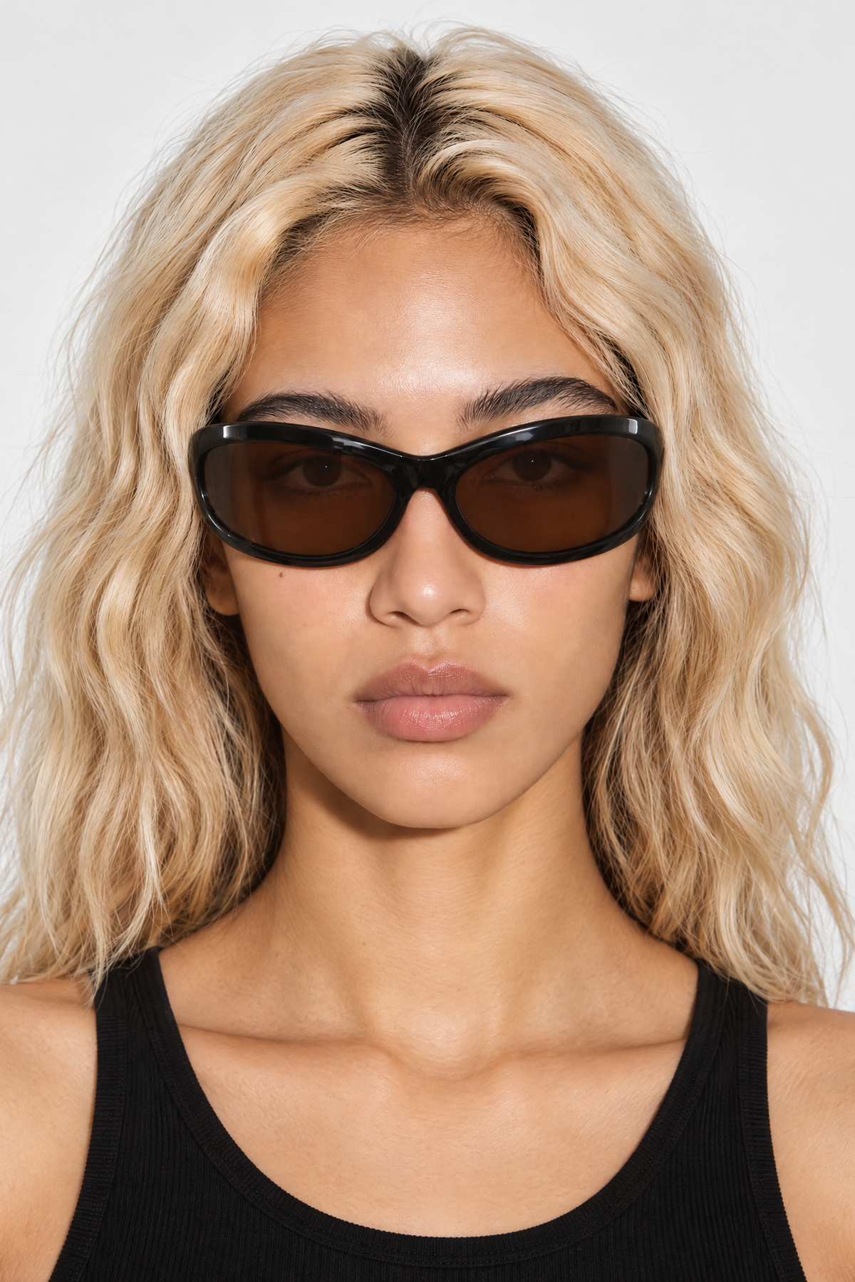 A close-up of a model with blonde wavy hair wearing black wrap around sunglasses with dark brown lenses and a black ribbed tank top, set against a plain light gray background.