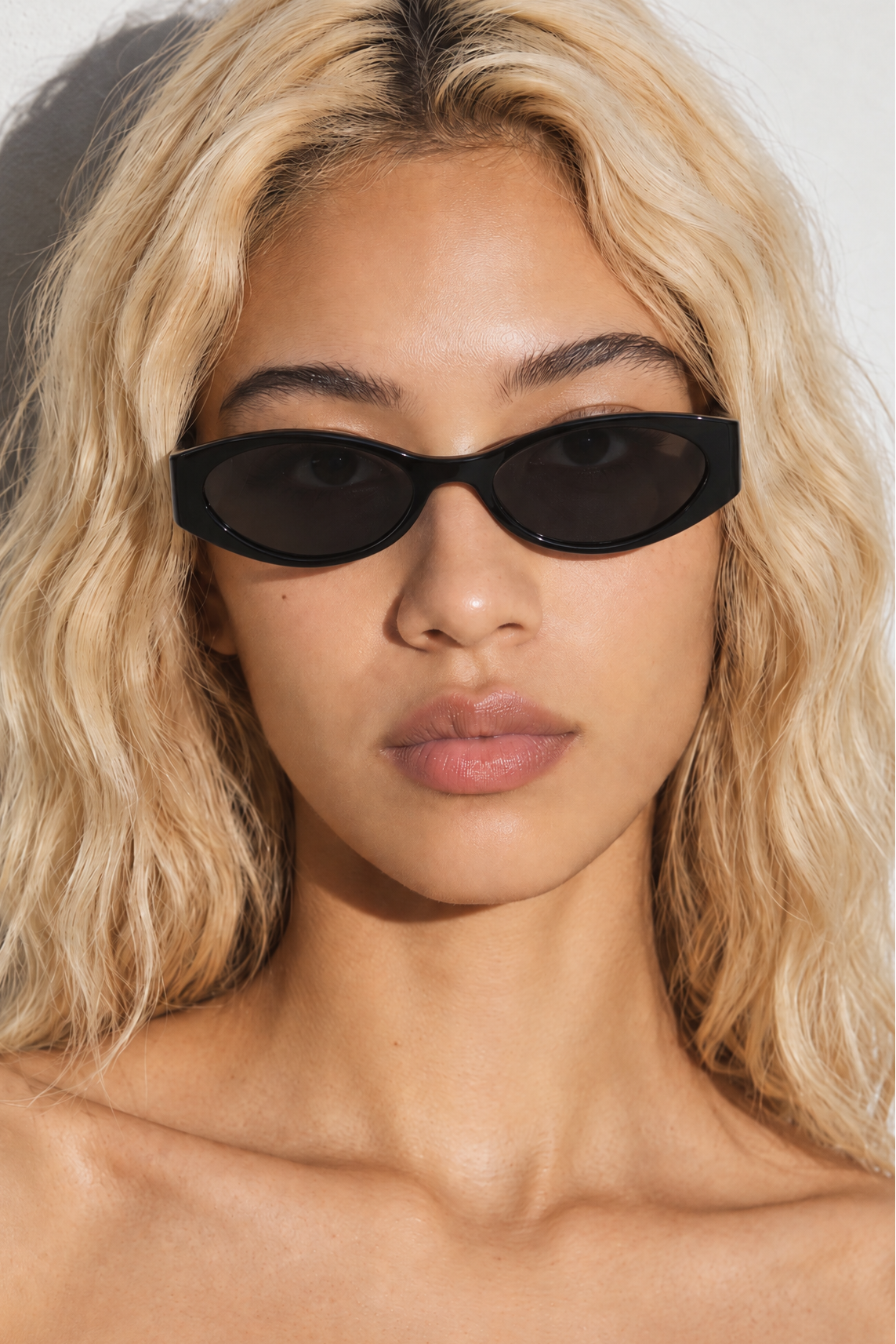 A close-up of a model with blonde wavy hair wearing black oval sunglasses against a light gray background.