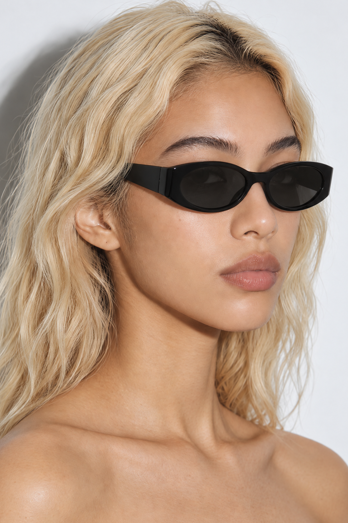 A close-up of a woman with blonde hair wearing black oval sunglasses against a plain light gray background.