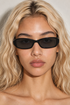 A close-up portrait of a model with wavy blonde hair wearing black rectangle sunglasses against a neutral light gray background.