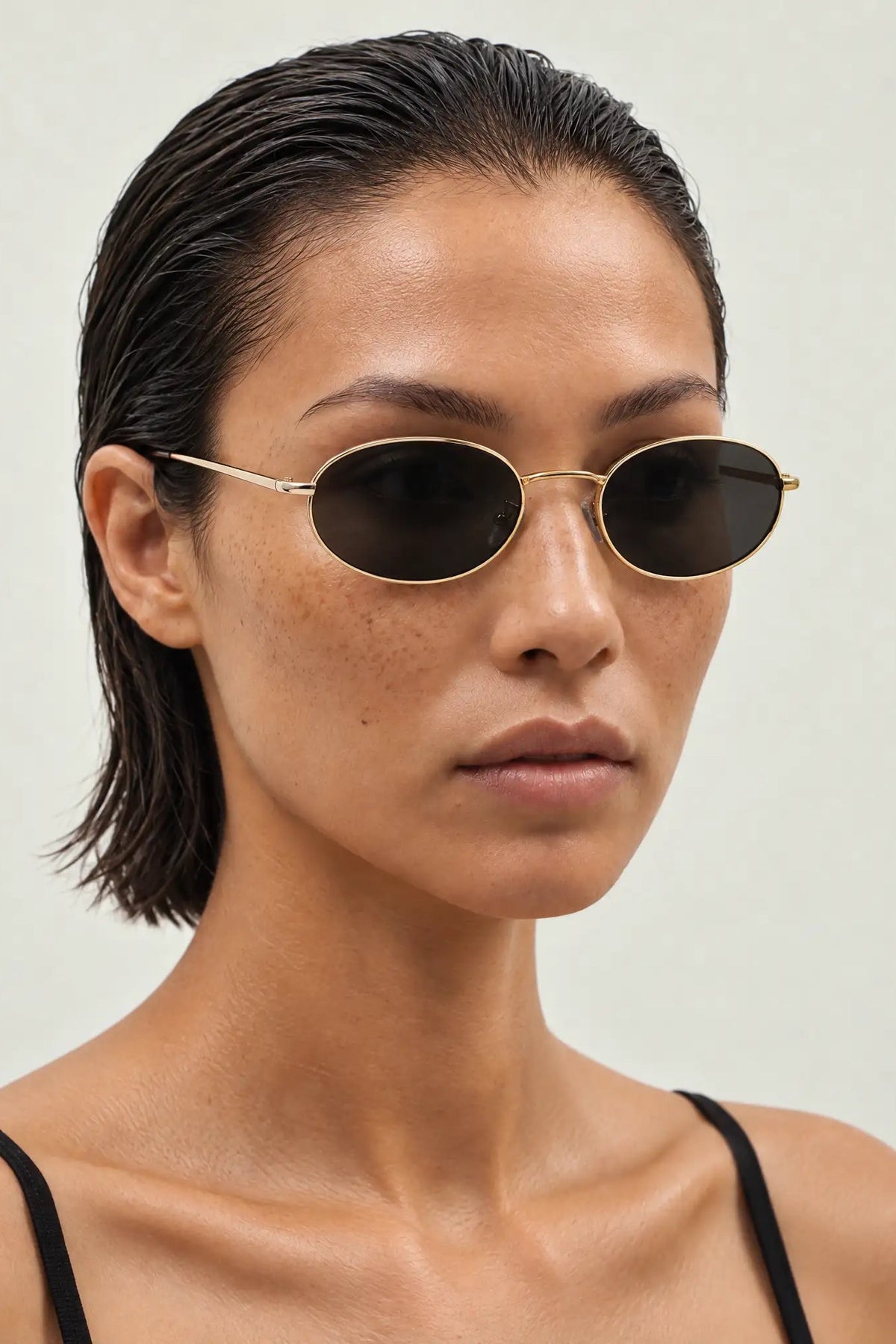 A close-up of a model wearing oval sunglasses with thin gold metal frames and dark lenses, set against a plain light gray background.