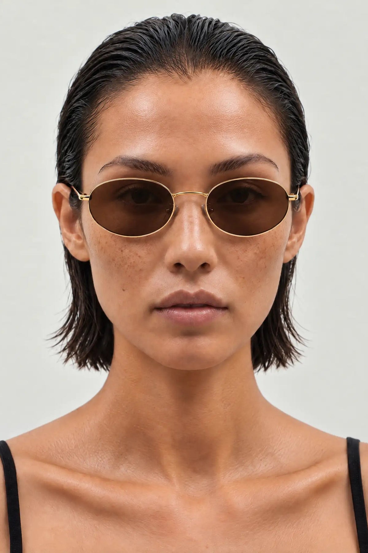 A close-up of a model with slicked-back dark hair wearing oval gold-framed sunglasses with dark brown lenses, set against a plain light gray background.