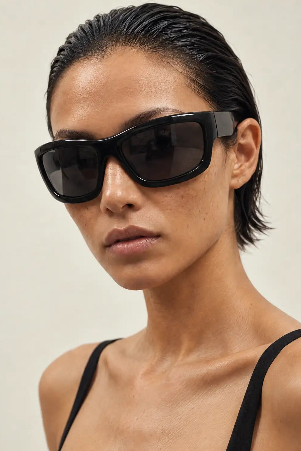A close-up of a model wearing thick black sunglasses with dark lenses and a black spaghetti-strap top against a plain light beige background.