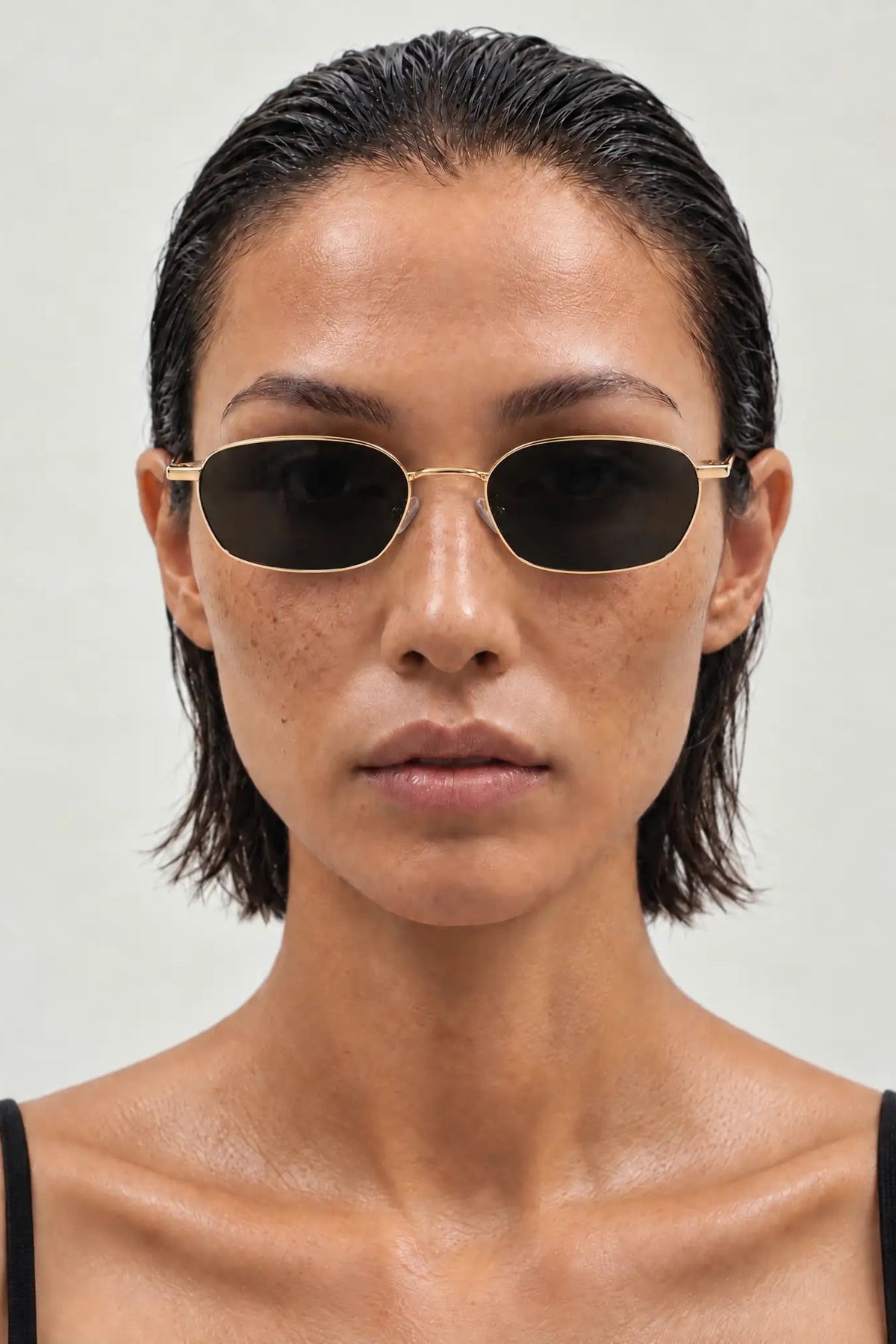 A close-up of a model wearing slim, oval-shaped sunglasses with a gold metal frame and dark lenses, set against a plain light gray background.