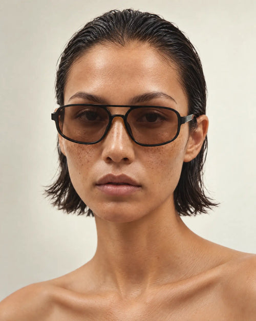 A close-up portrait of a model wearing aviator sunglasses with black frame and brown tinted lenses, set against a plain light beige background.
