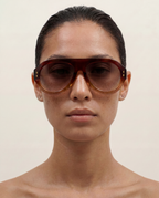 A close-up portrait of a model wearing oversized aviator-style sunglasses with a translucent dark brown frame and tinted lenses, set against a plain light beige background.