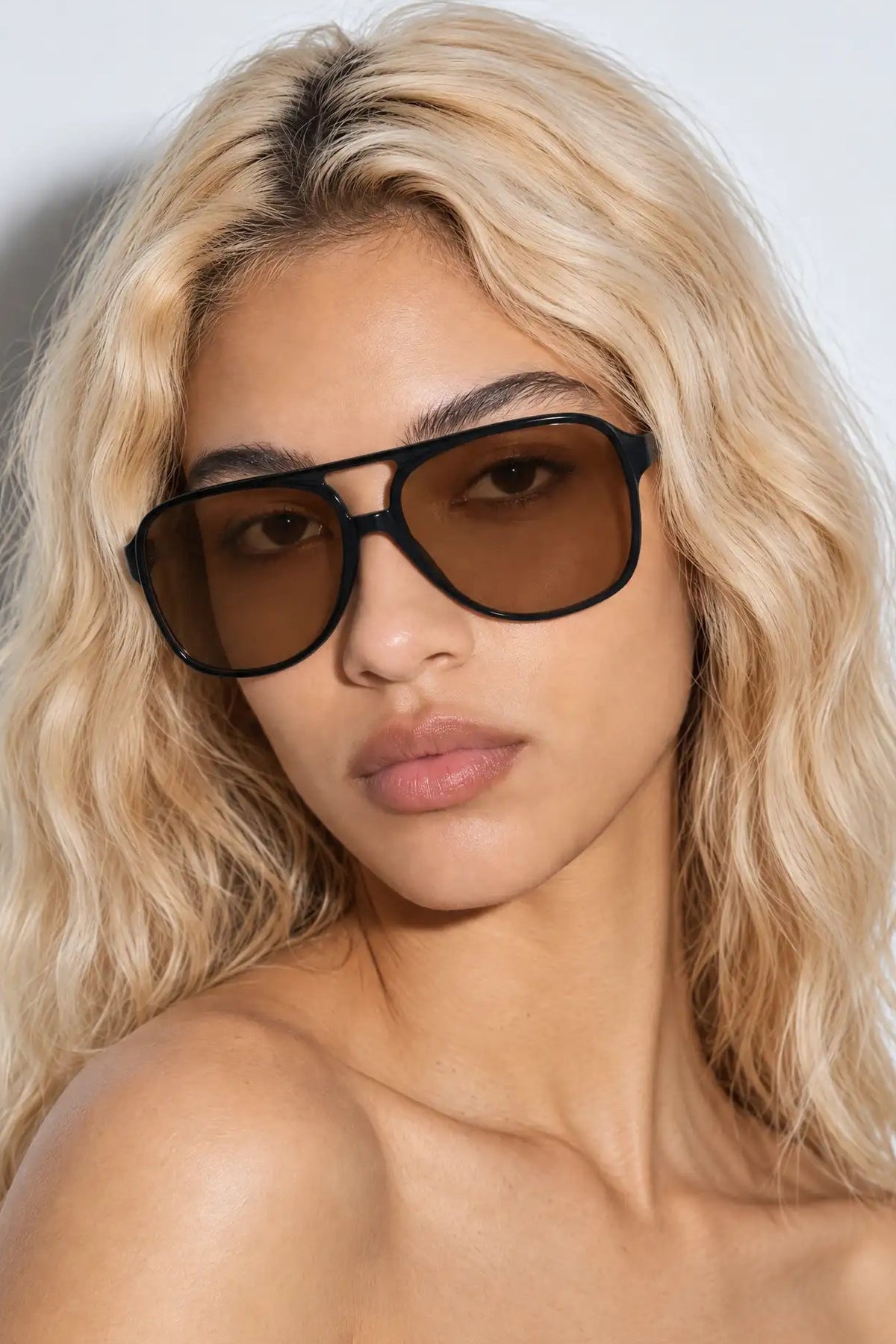 A close-up of a woman with blonde wavy hair wearing oversized aviator sunglasses with thick black frames and dark brown lenses, set against a plain light gray background.