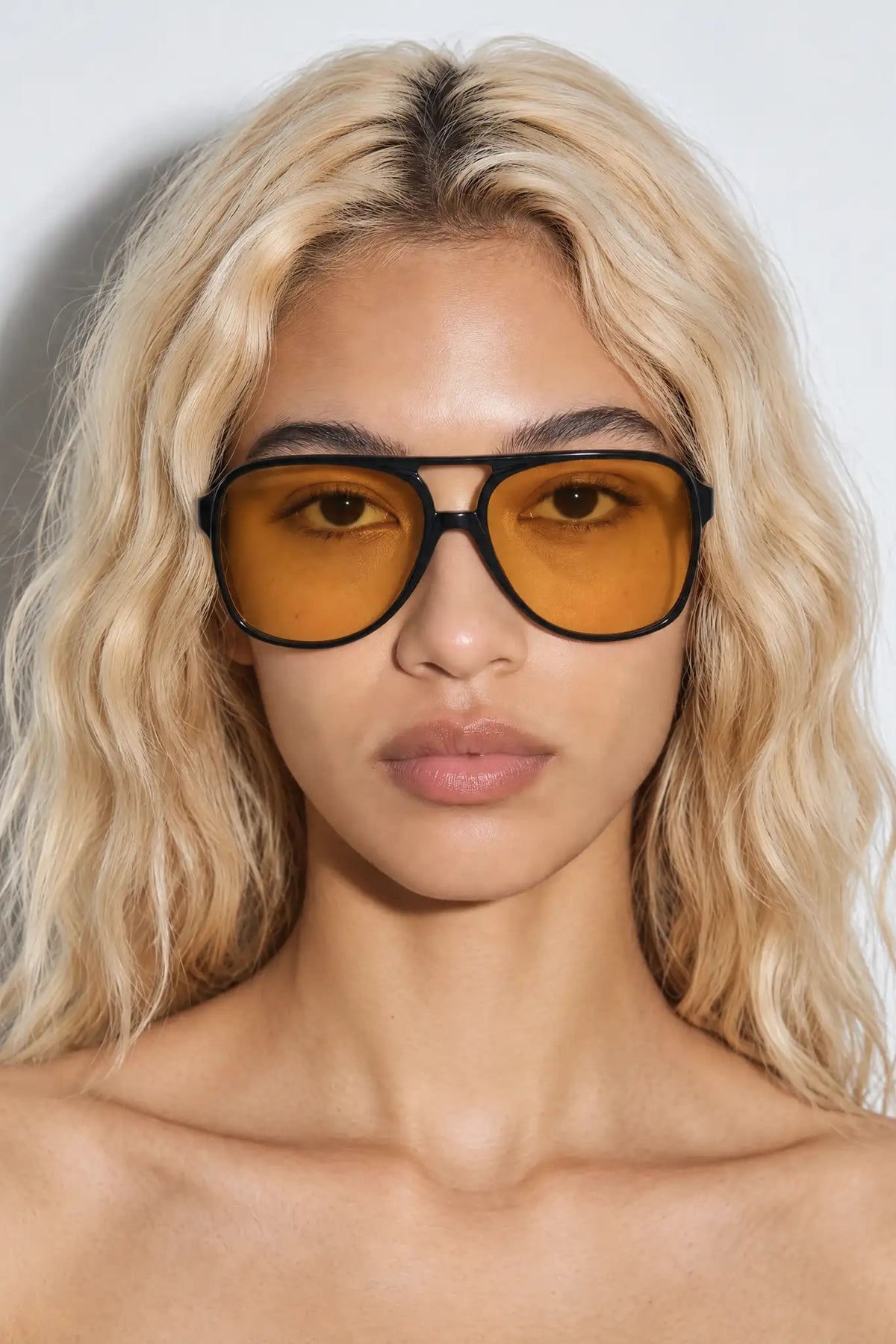 A close-up of a model with blonde wavy hair wearing oversized aviator sunglasses with thick black frames and amber-yellow lenses, set against a plain light gray background.