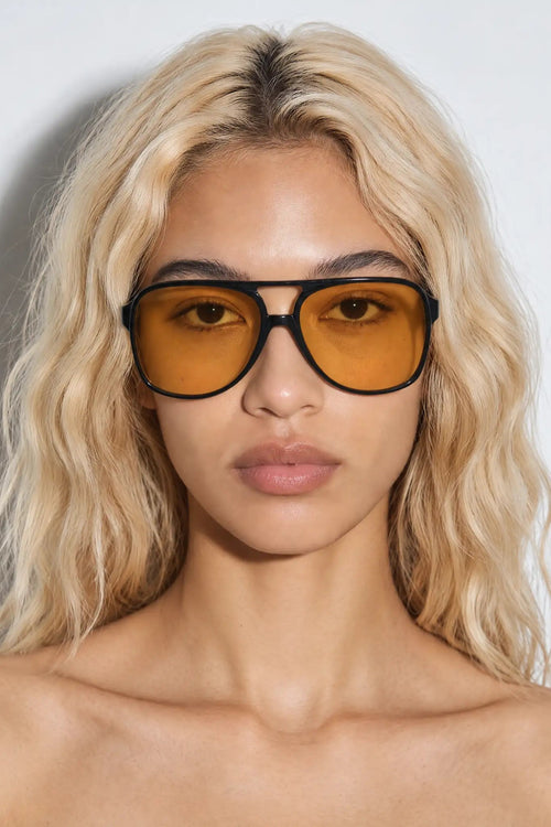 A close-up of a model with blonde wavy hair wearing oversized aviator sunglasses with thick black frames and amber-yellow lenses, set against a plain light gray background.