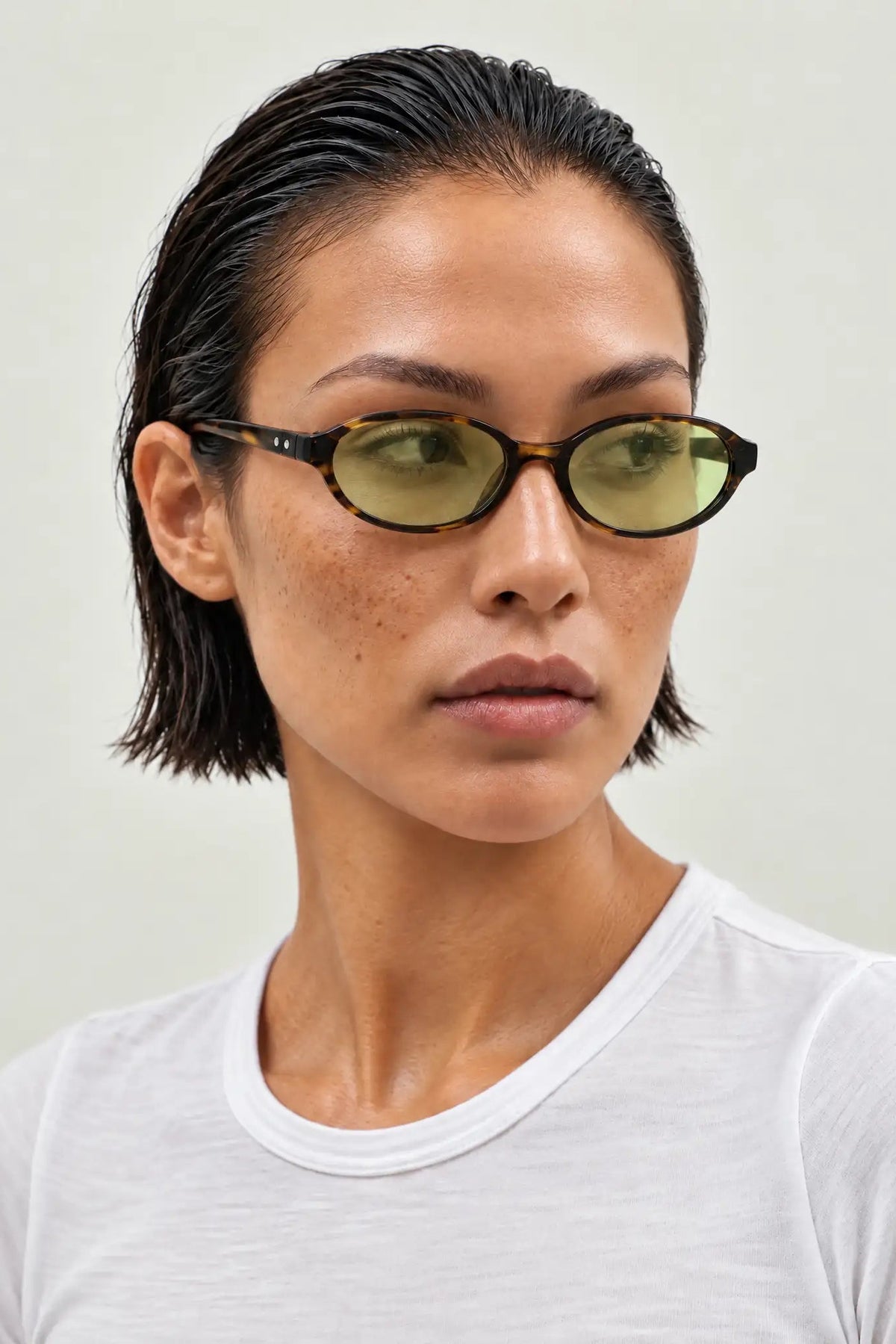 A close-up of a model with short dark hair wearing cat-eye sunglasses with olive green tinted lenses and dark tortoiseshell frames, paired with a white t-shirt against a light gray background.