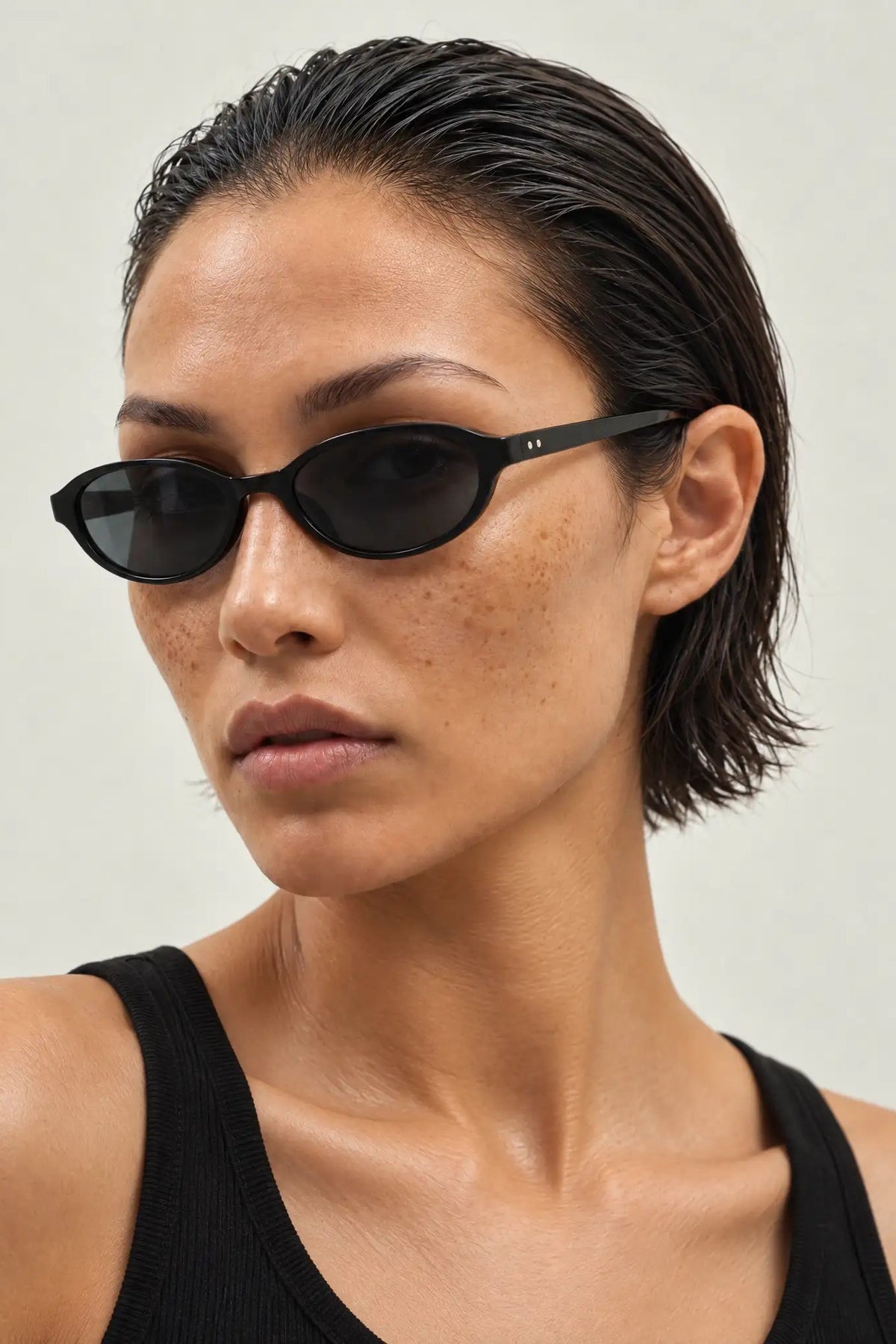 A close-up of a model with slicked-back dark hair wearing slim black cat-eye sunglasses and a black ribbed tank top against a light gray background.